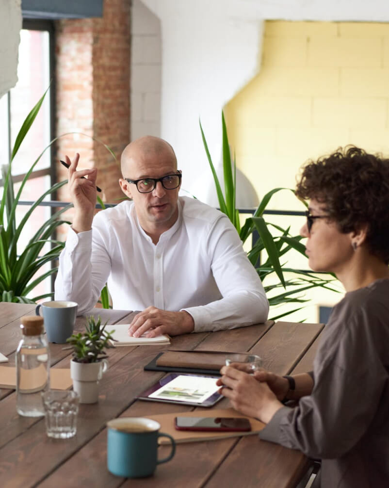 Man and woman having a discussion at a wooden table with coffee and plants