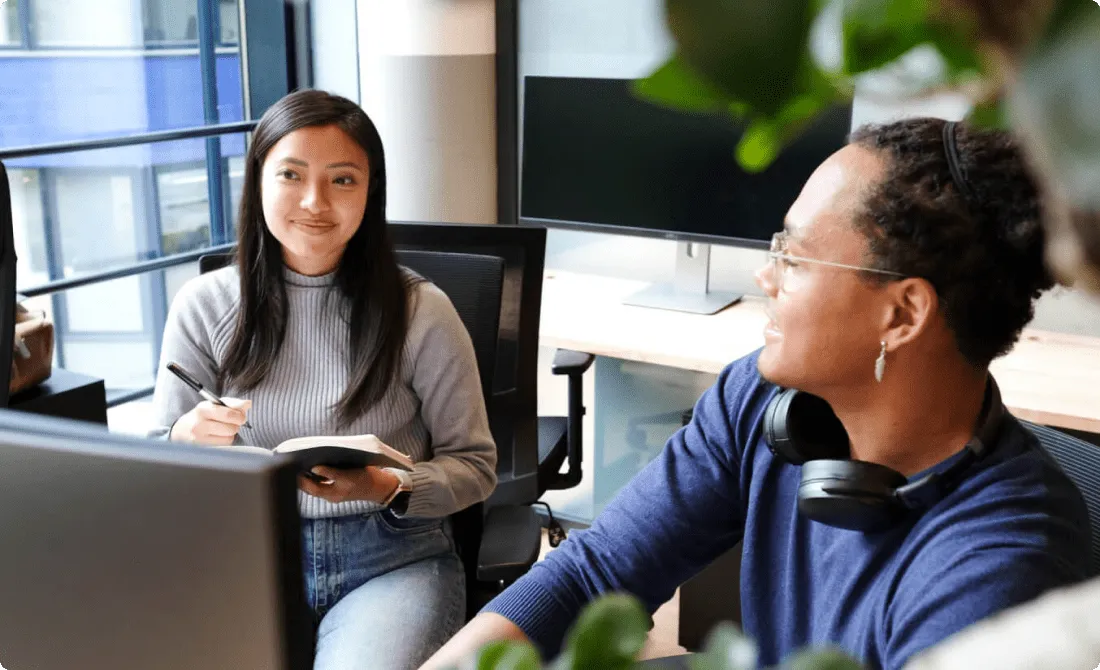 Two coworkers sitting at a modern office desk having a friendly conversation. One person is holding a notebook and pen, while the other wears headphones around the neck and smiles. A large monitor is visible in the background with natural light coming through the window.
