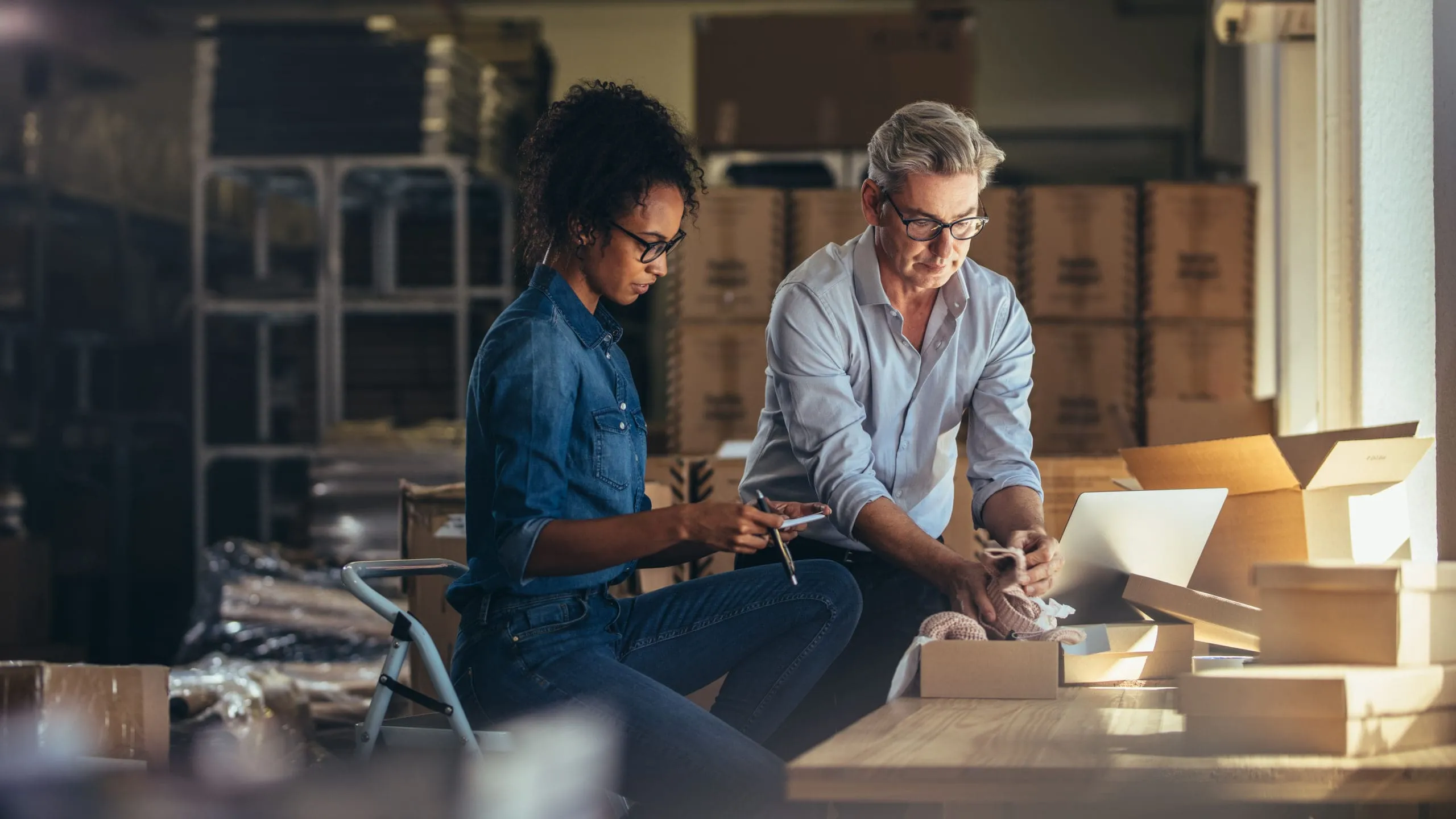 Two people working together in a warehouse, packaging products into cardboard boxes with a laptop on the table.