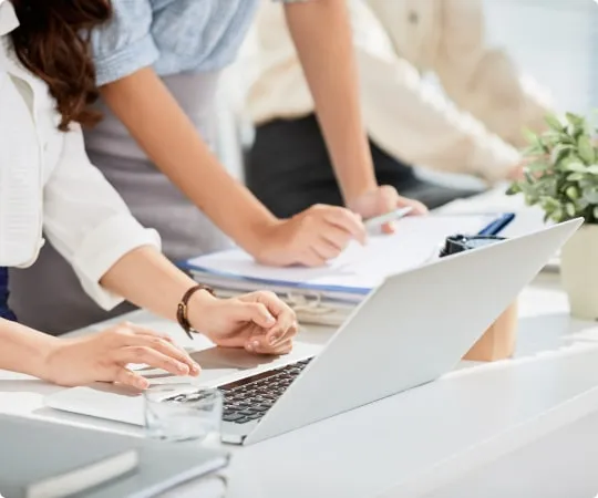 Close-up of business professionals working at a desk with a laptop, documents, and a clipboard, collaborating in a bright office setting.