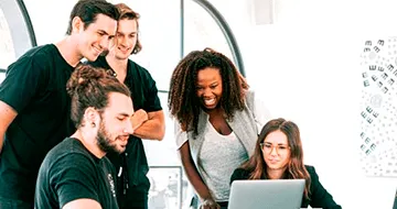 Five people gather around a laptop in a bright office, looking at the screen and smiling while working together.