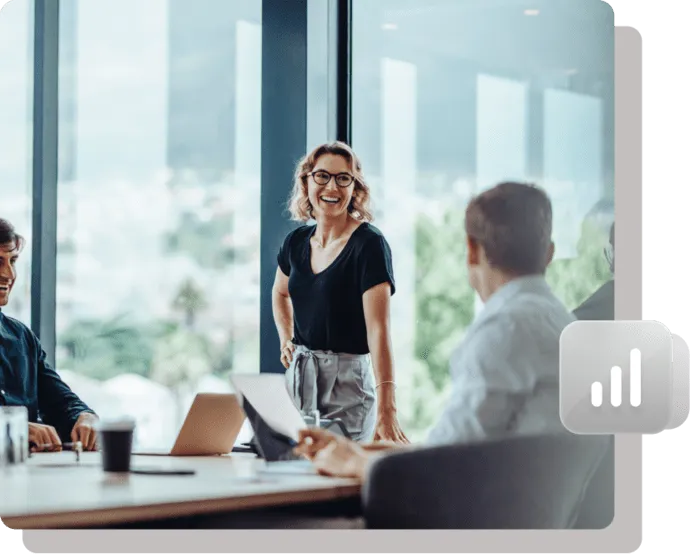 Smiling businesswoman presenting in a modern office meeting with colleagues around a conference table and laptops.