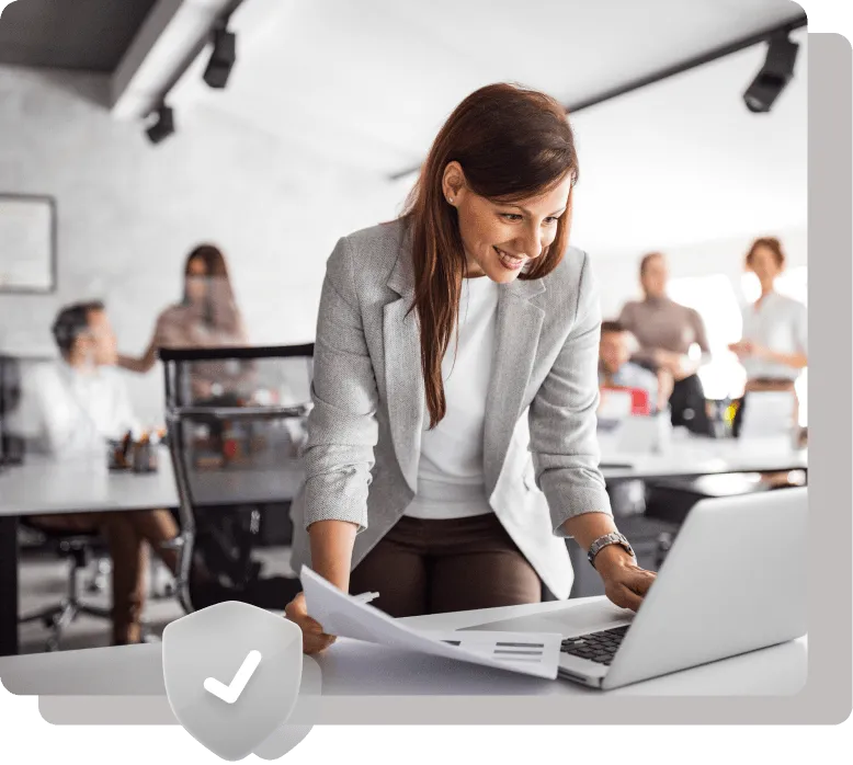 Smiling businesswoman in a modern office leaning over a desk while working on a laptop, holding documents.