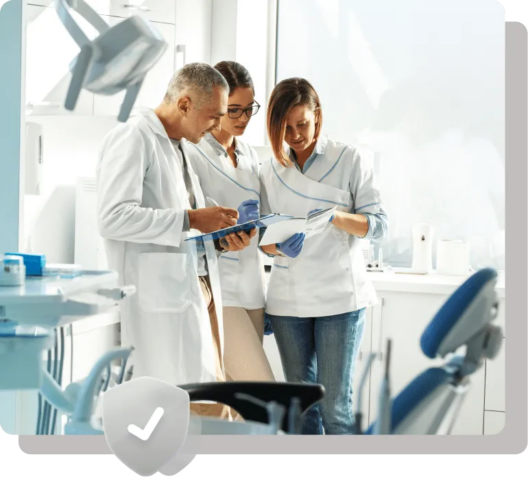 Three dental professionals in a modern clinic discussing notes and reviewing patient files. They are wearing white lab coats and gloves, standing beside dental chairs and equipment.