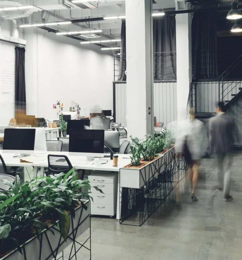 Modern open-concept office with white desks, black computer monitors, ergonomic chairs, and indoor plants arranged in rows. Several people are walking through the space, captured with motion blur, creating a sense of activity.