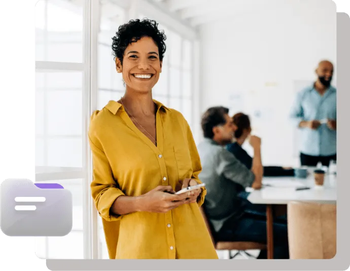 Confident woman in yellow shirt smiling while holding a phone, with a team meeting taking place in the background.