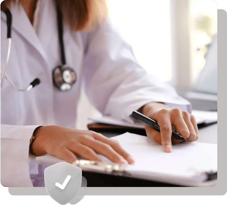 Close-up of a healthcare professional in a white lab coat writing on a clipboard, with a stethoscope around their neck and a laptop in the background.