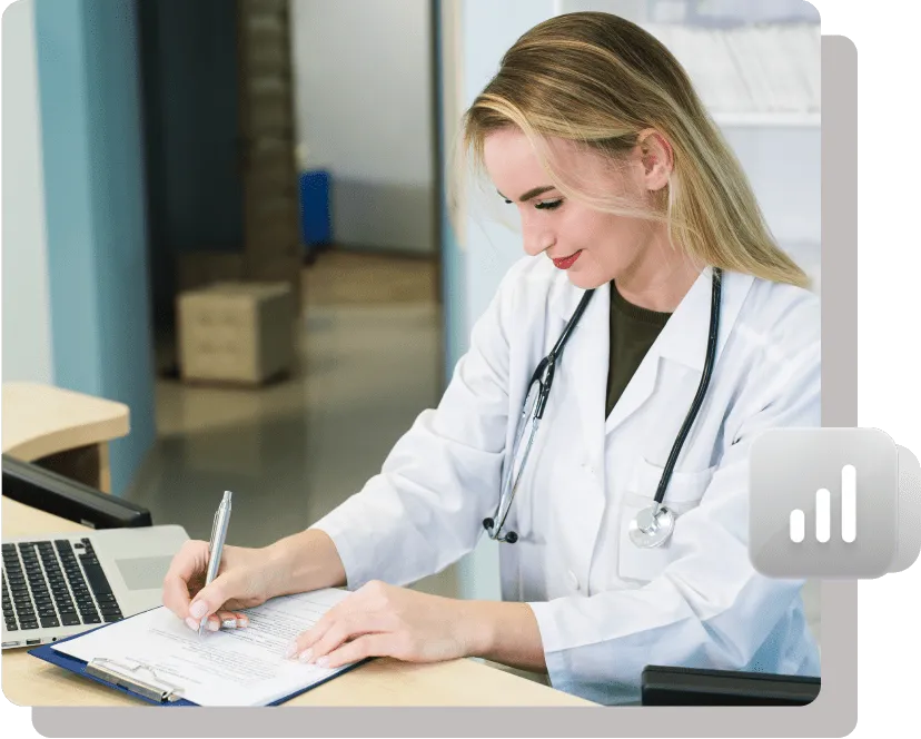 Female doctor in a white coat sitting at a desk, writing on a clipboard with a pen, with a laptop beside her. She is smiling and appears focused.