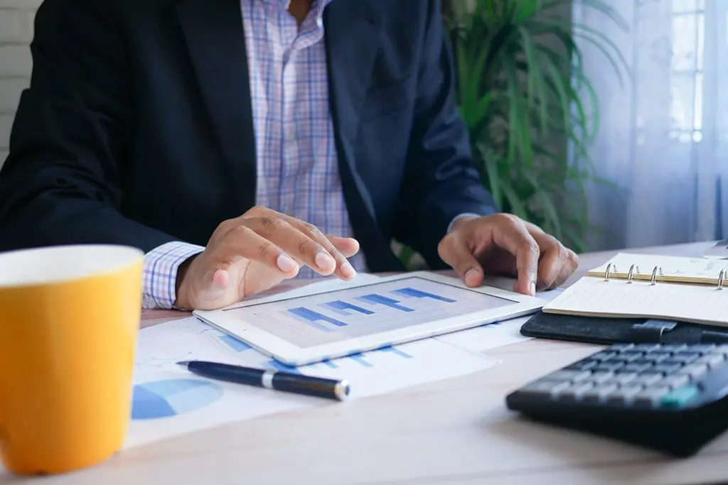 Businessman analyzing bar charts on a tablet with documents, notebook, and calculator on desk.