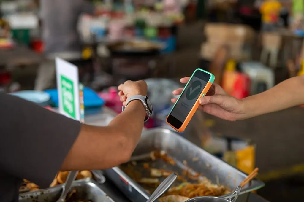 Customer using smartwatch for contactless payment at a street food stall, confirmed by a mobile phone screen with a green checkmark.