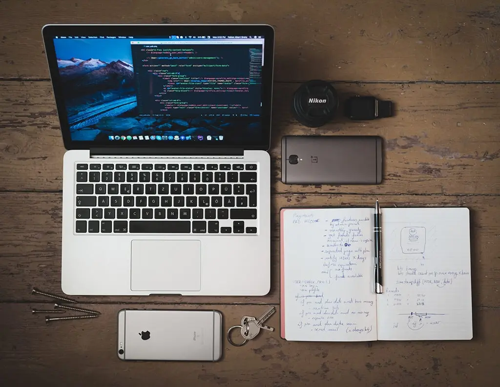 Top view of a developer’s workspace with a MacBook showing code, smartphones, handwritten notes, and accessories on a wooden desk.