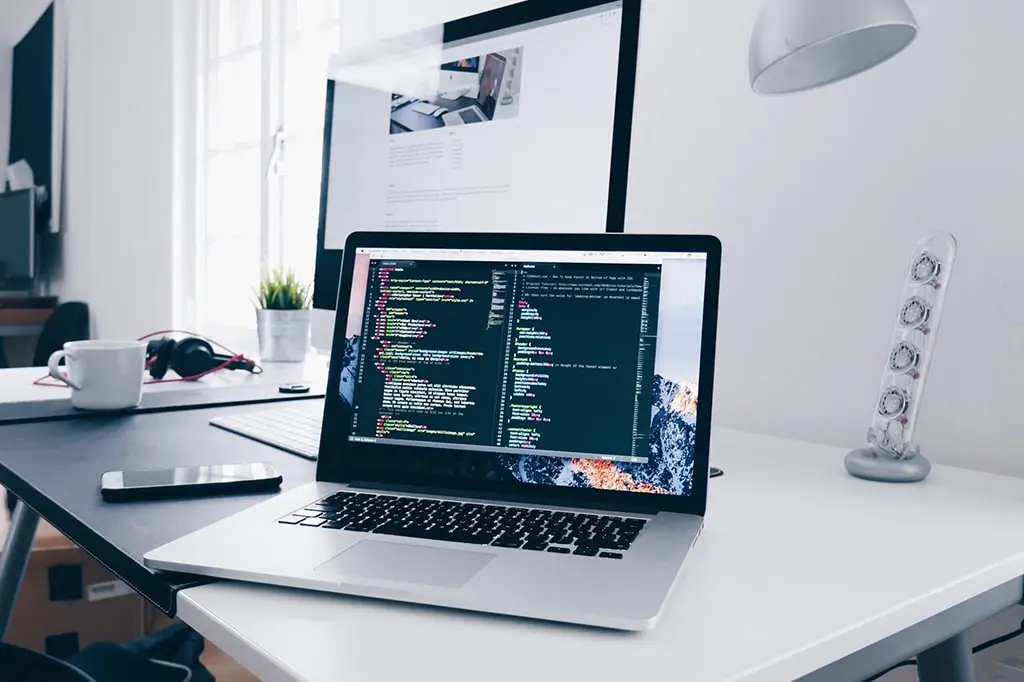 Modern workspace with a MacBook displaying code, a large external monitor in the background, a smartphone, headphones, and a coffee cup on the desk.