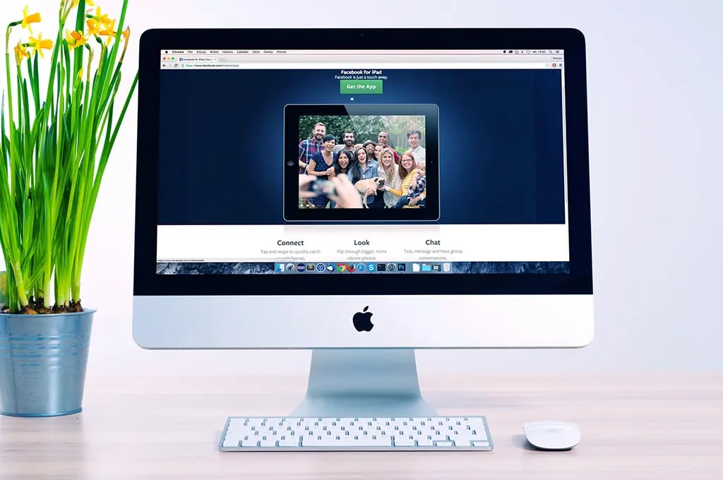 iMac displaying a Facebook for iPad promotional page, with a group photo on the screen; yellow daffodils in a metal pot on the left and a wireless keyboard and mouse on the wooden desk.