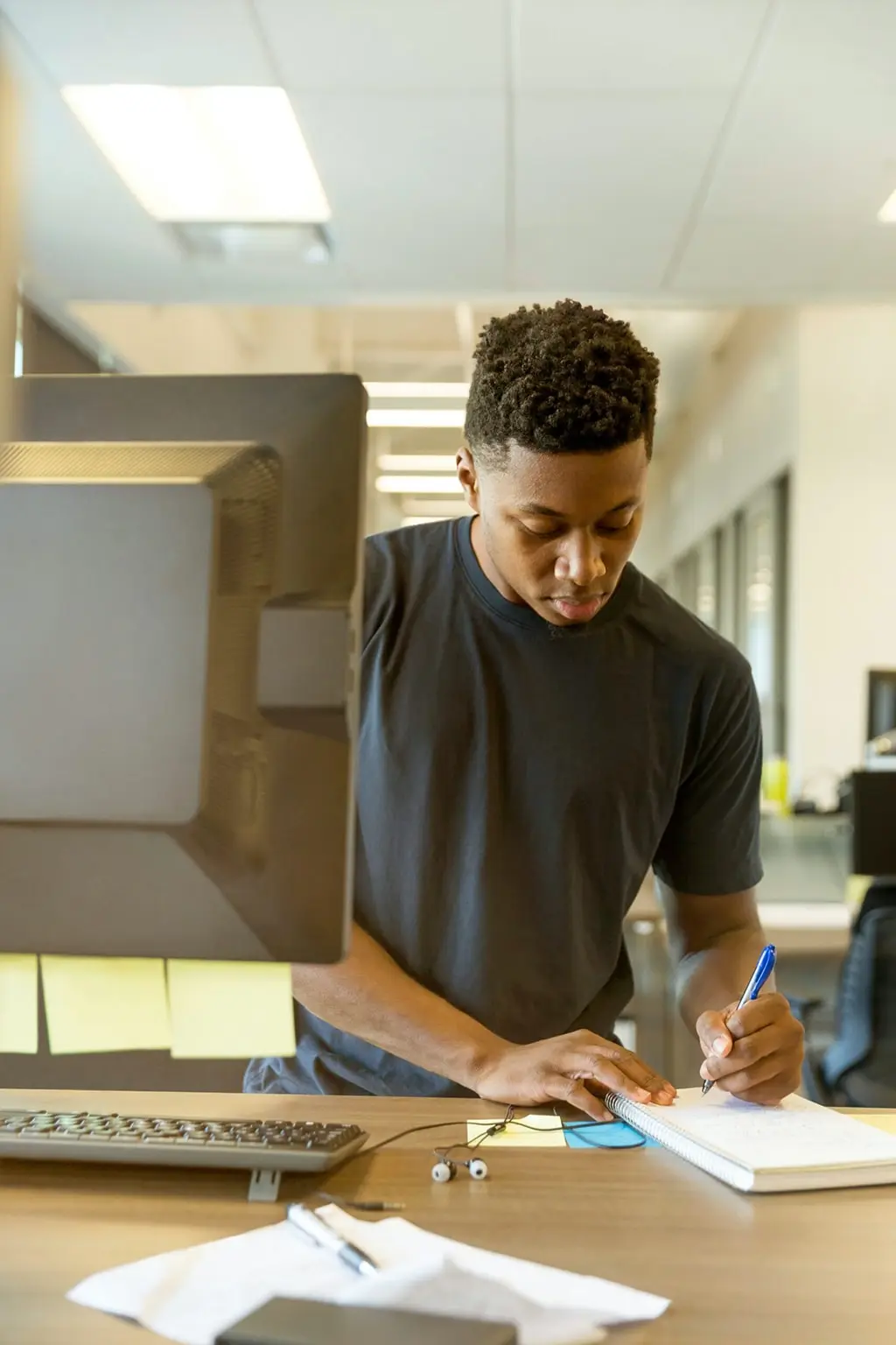 Man in black t-shirt writing in a notebook while working at a desk with a computer monitor in a modern office.
