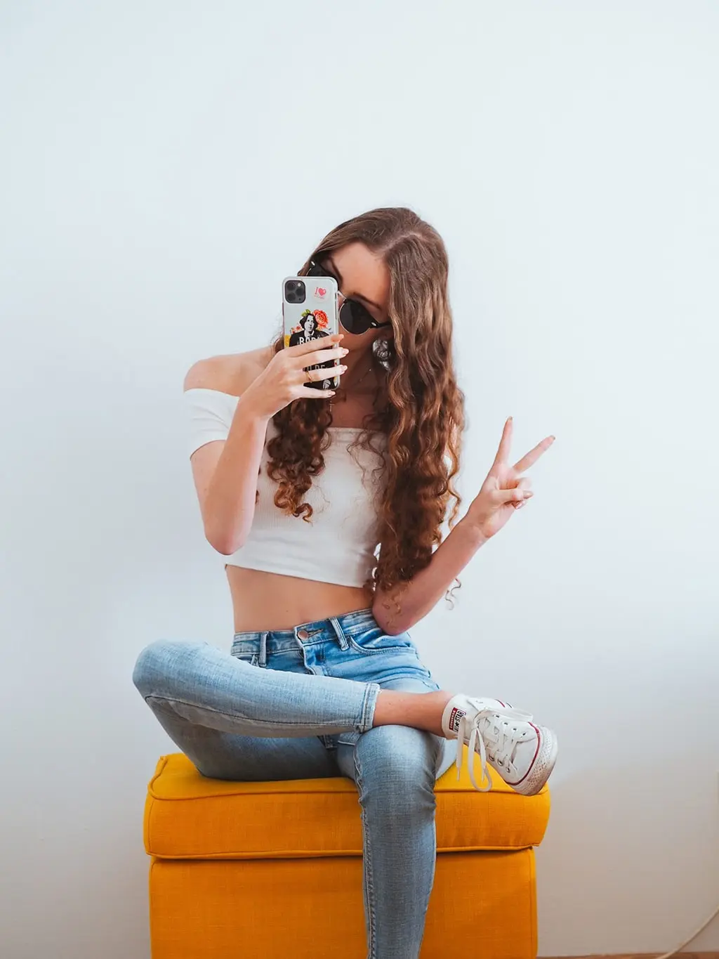 Young woman with long curly hair taking a selfie while sitting on a yellow stool, wearing sunglasses, white crop top, and jeans, posing with a peace sign.