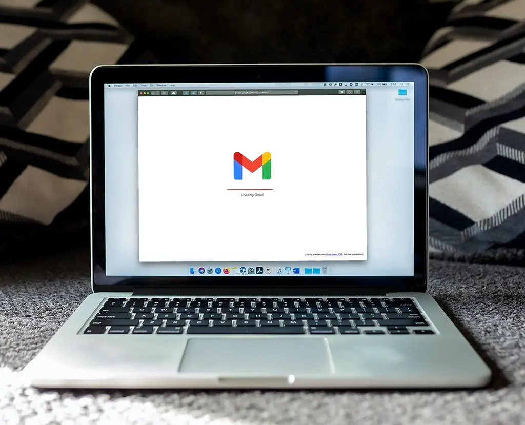 Laptop showing Gmail loading screen placed on carpet with pillows in background.