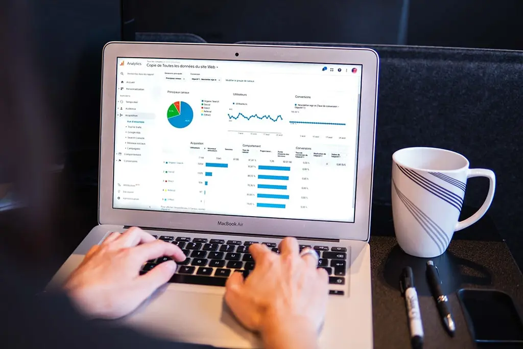 Person working on a MacBook Air displaying a Google Analytics dashboard with graphs and pie charts; a white coffee mug and markers rest on the black table beside the laptop.