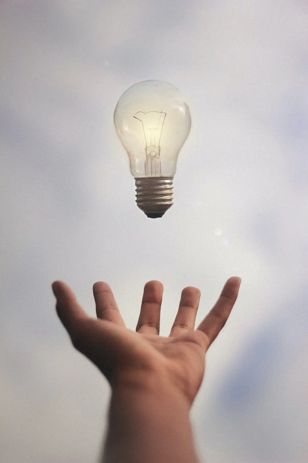 Outstretched hand reaching toward a floating light bulb against a cloudy sky, symbolizing creativity or inspiration.