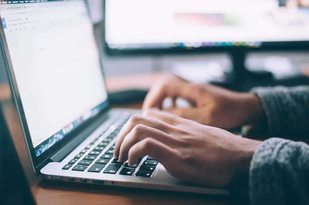 Close-up of hands typing on a laptop keyboard with a blurred monitor in the background.