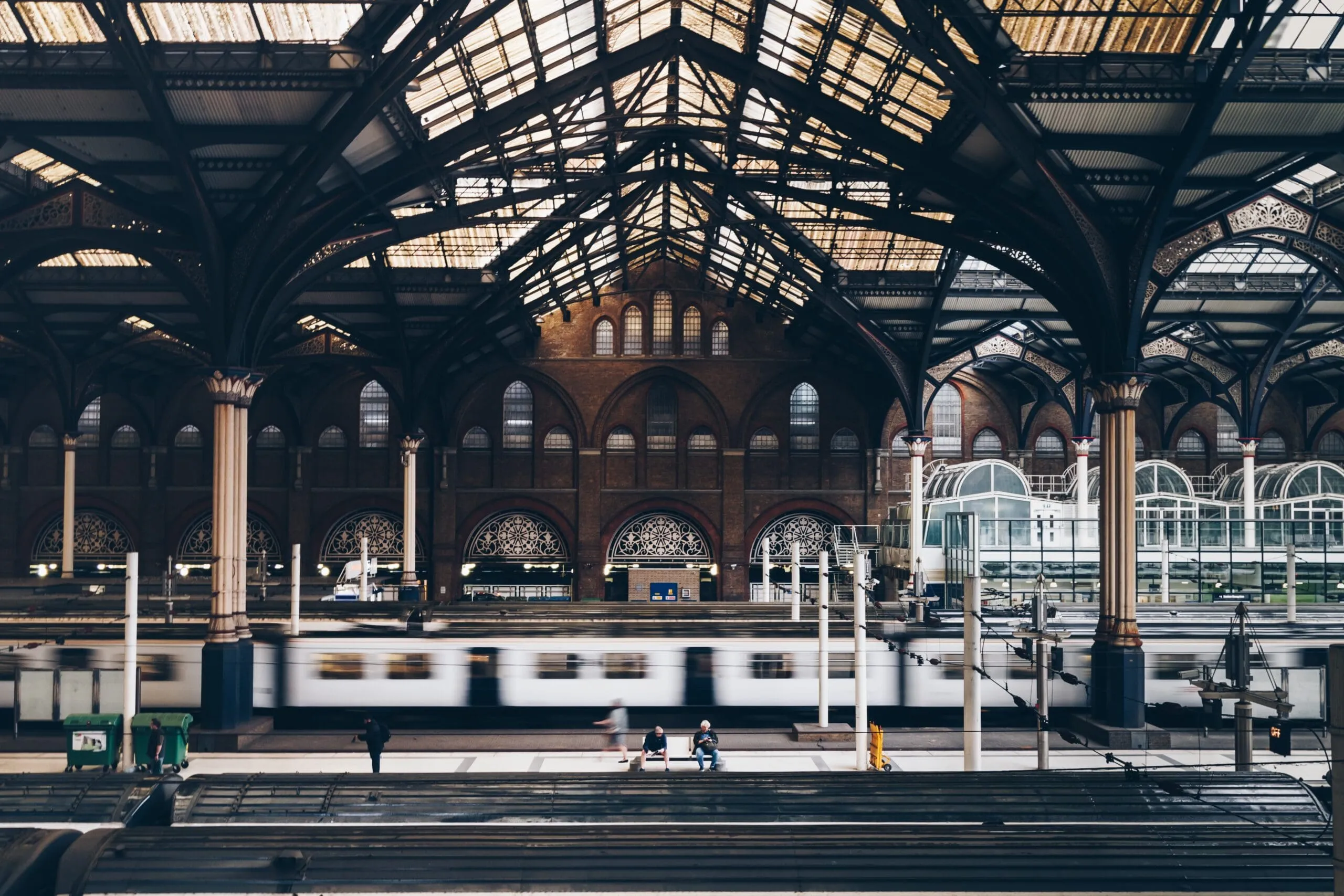 A train moves through a large, ornate railway station with high arched windows, columns, and a glass-paneled roof; a few people stand on the platform.