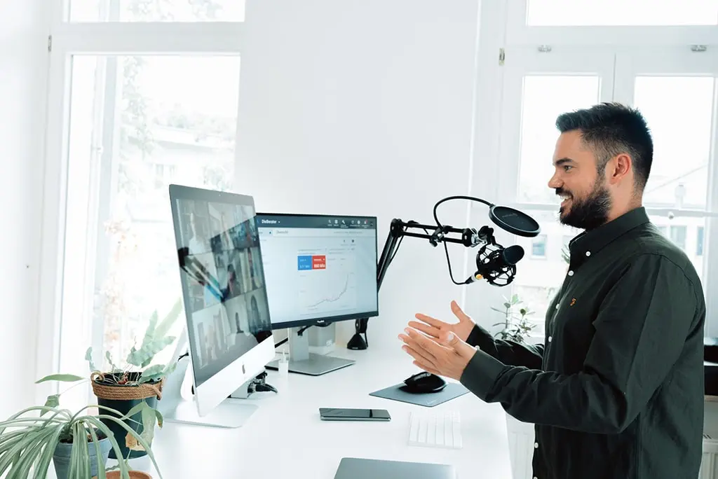 Smiling man standing at a desk with microphone, delivering an online presentation with charts and video call participants on dual monitors.