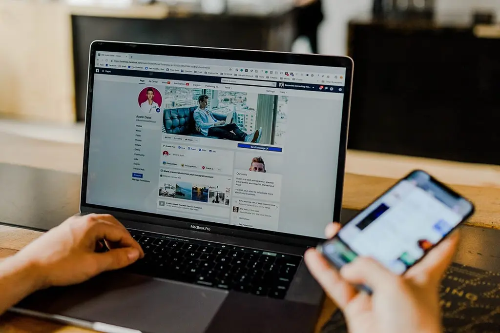Person managing social media on a MacBook and smartphone simultaneously.