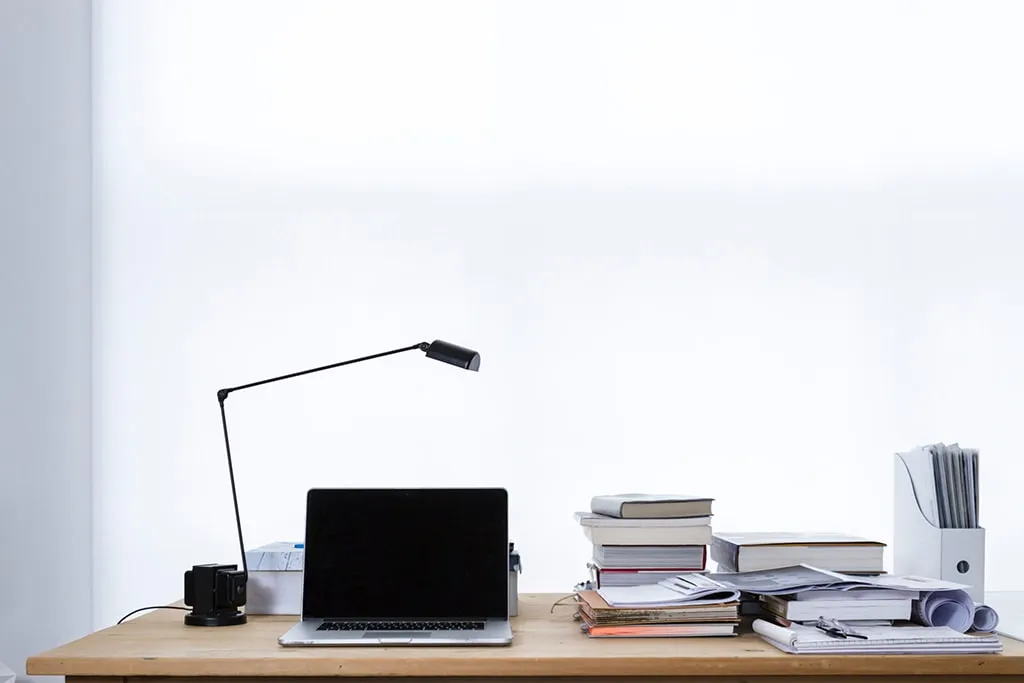 A wooden desk with a closed laptop, desk lamp, coffee mug, stacked books, papers, and a file organizer in front of a bright window.