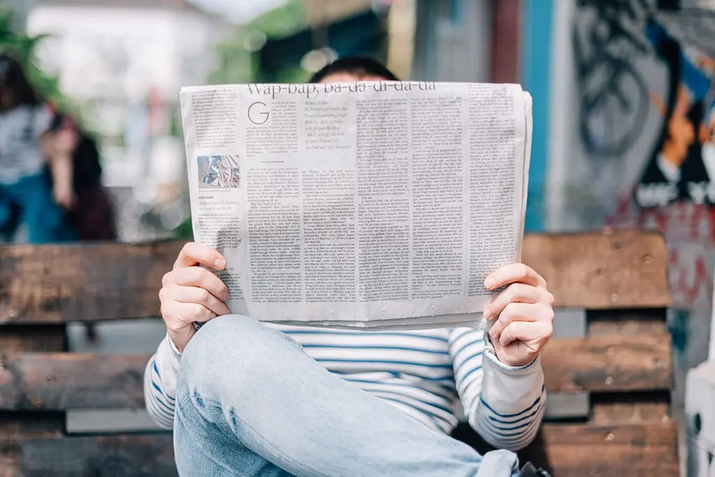 Man in striped shirt reading a newspaper while sitting on a wooden bench outdoors.