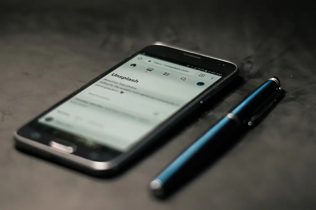 Smartphone displaying the Unsplash website placed on a dark surface beside a blue metallic pen.