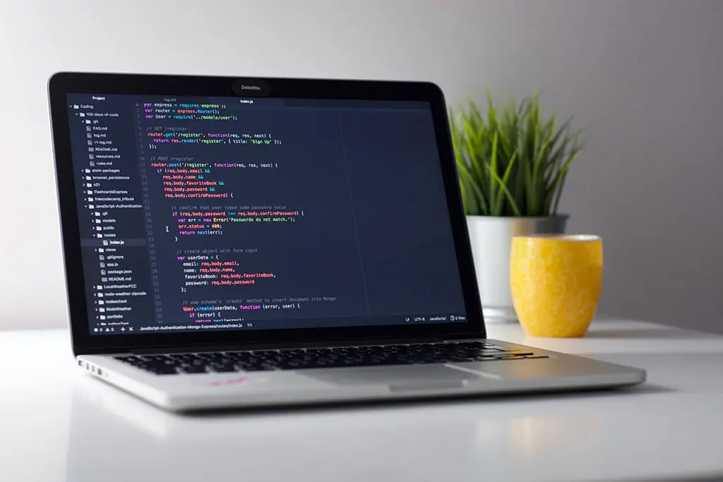 Open laptop displaying JavaScript code in a dark-themed editor, placed on a white desk with a yellow mug and a green plant in the background.