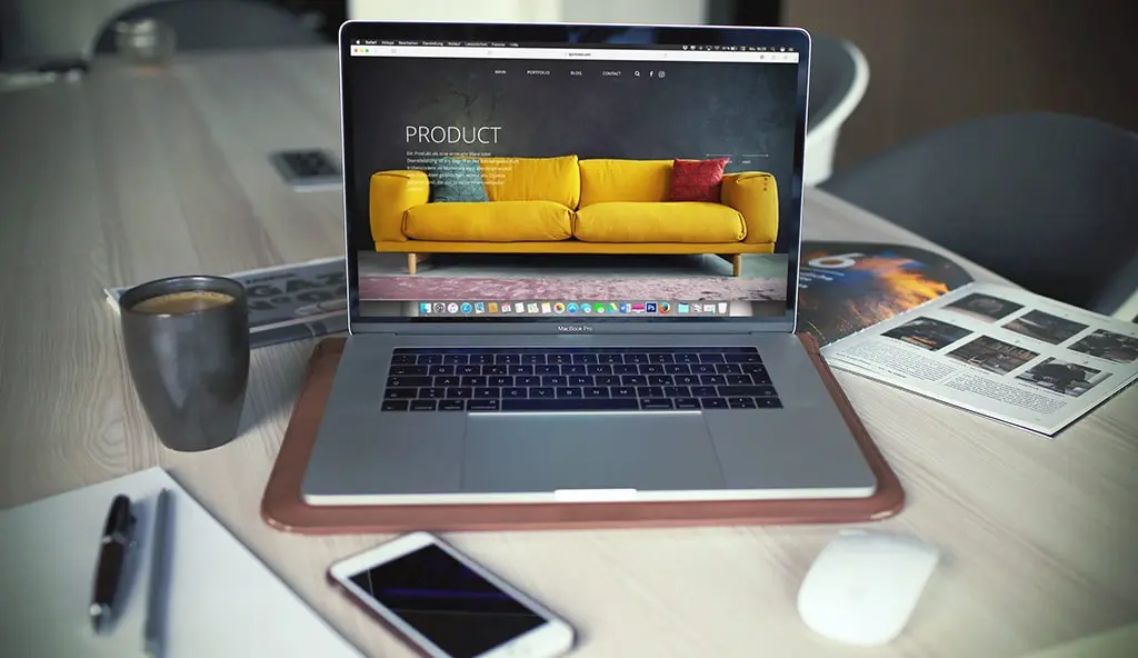 MacBook displaying a product website featuring a yellow sofa, placed on a desk with coffee, a phone, and magazines.