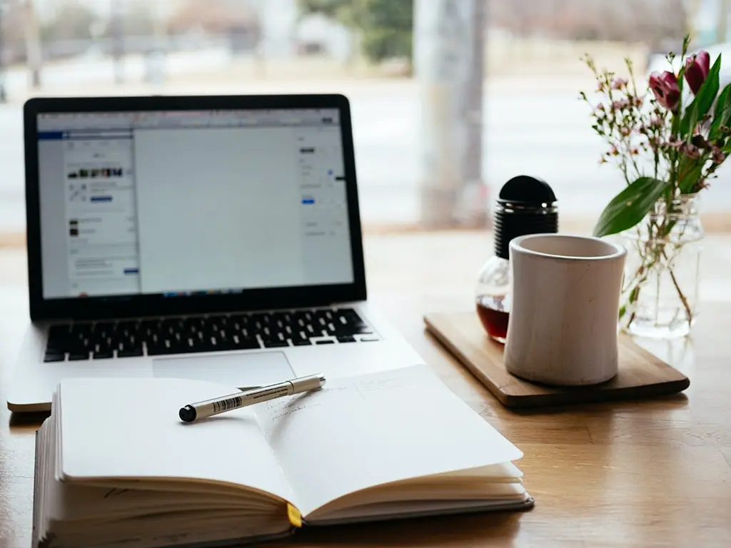 Open laptop with a blank document, a notebook with a pen, coffee mug, and vase with flowers on a wooden desk near a window.
