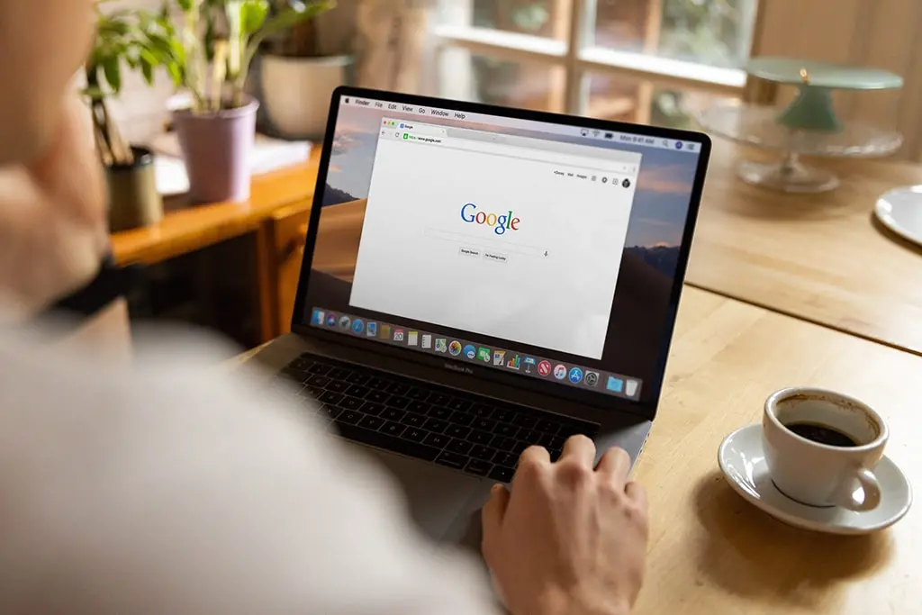 Person using a MacBook with Google open on the screen, sitting at a wooden table with a cup of coffee.