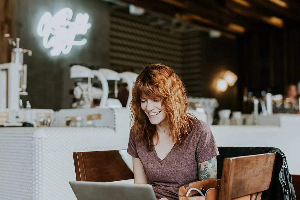 Happy woman with wavy red hair using a laptop at a cozy coffee shop.