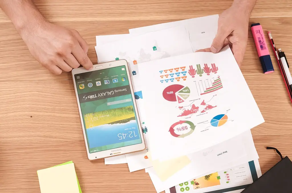 Person analyzing colorful infographics on paper with a Samsung Galaxy Tab S placed on a wooden desk.
