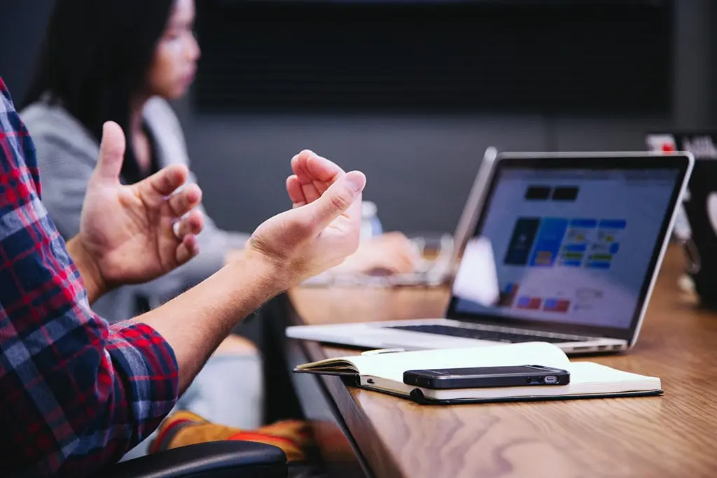 Close-up of a person gesturing during a team discussion with a laptop and notebook on the table.