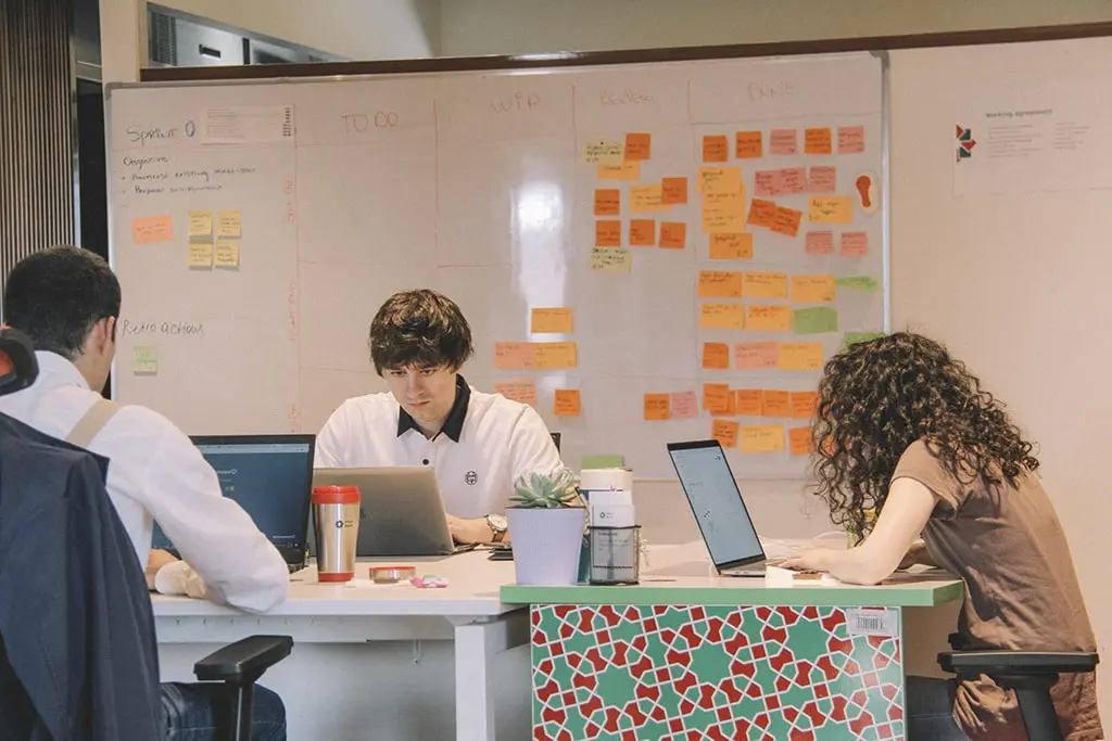 Team of three people working on laptops in front of an agile sprint board with colorful sticky notes.