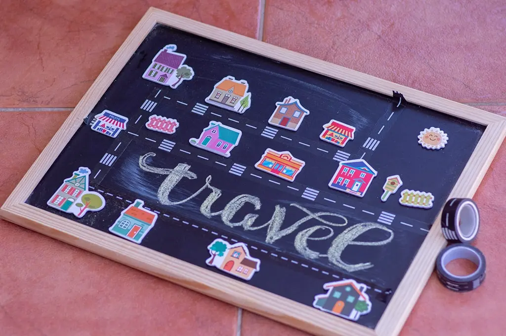 Travel-themed blackboard with sticker illustrations of houses, roads, and landmarks arranged on a tiled floor