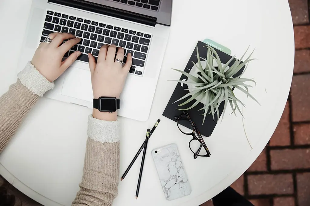 Top view of a person typing on a laptop with accessories like a plant, phone, notebook, and glasses on a white desk.