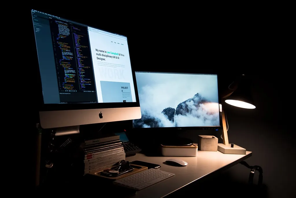 Dual monitor desk setup with coding and design interface on screen, lit by a desk lamp in a dark room.
