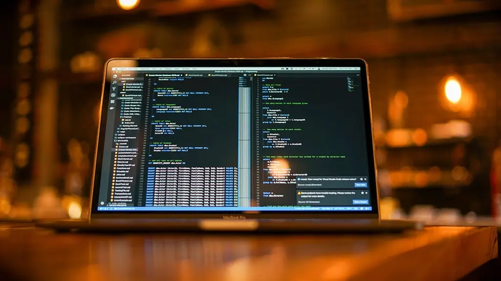 Laptop screen displaying SQL code in Visual Studio Code with a blurred background of a cozy, dimly lit room.