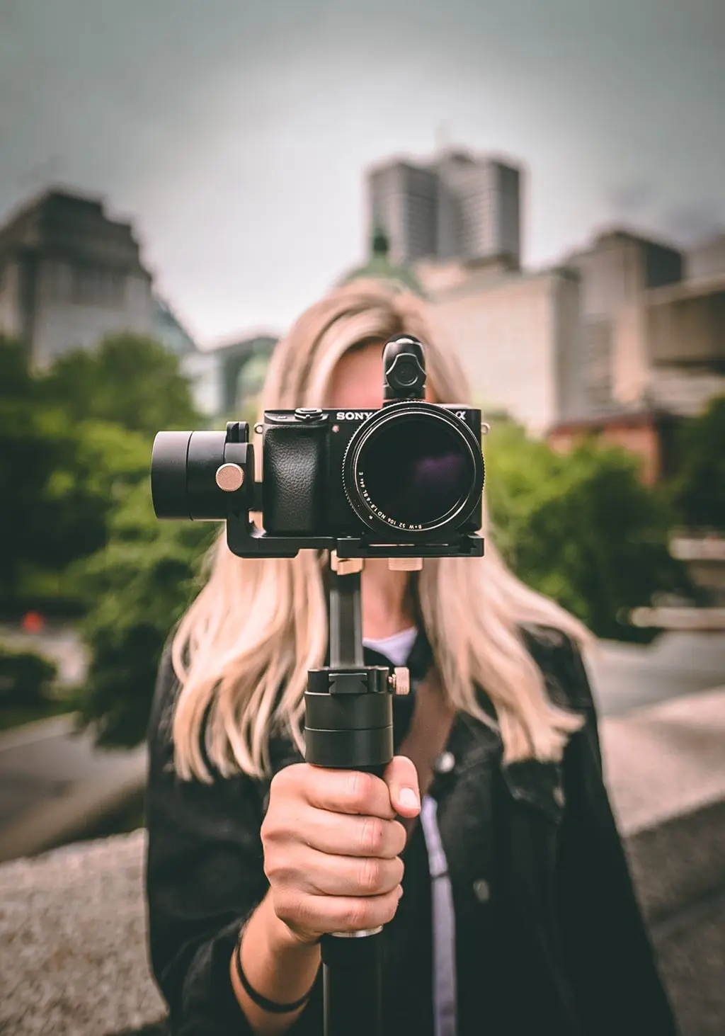 Blonde woman holding a Sony camera mounted on a gimbal, with an urban cityscape in the background.