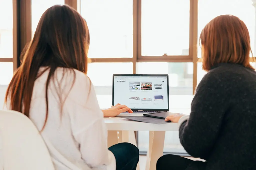 Two women sitting at a table reviewing products on a laptop screen in a bright office setting.