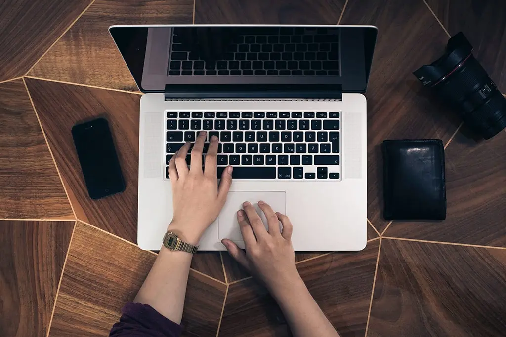 Top view of hands typing on a laptop keyboard with a smartphone, camera lens, and wallet placed on a wooden floor.