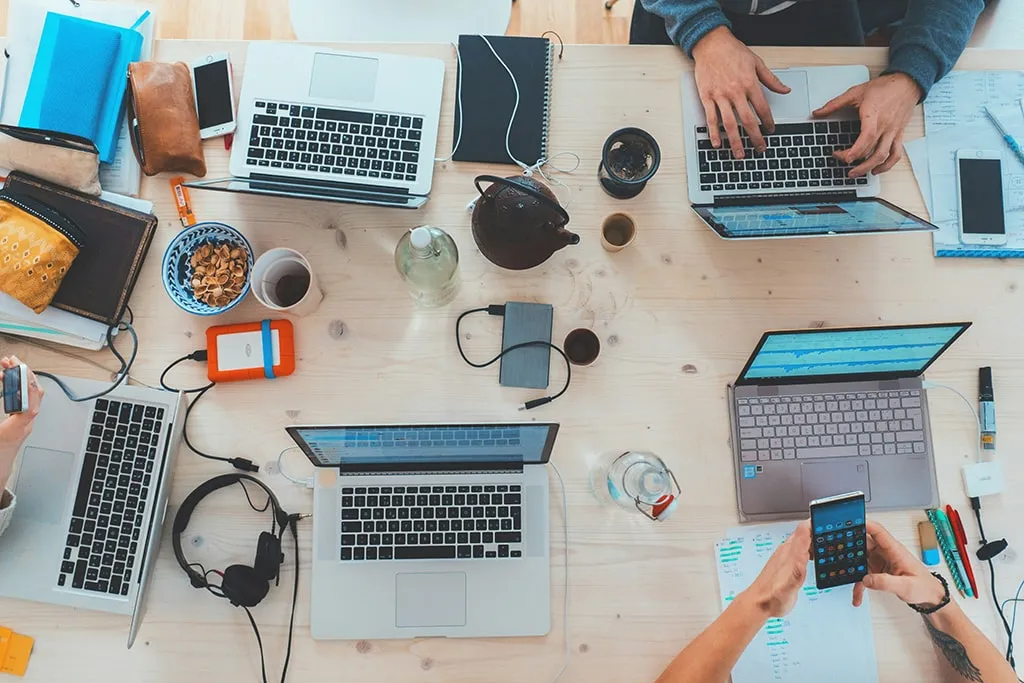 Overhead view of a shared coworking desk with multiple laptops, notebooks, smartphones, and drinks, used by a creative team collaborating