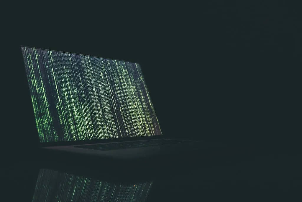 Laptop in a dark room displaying green matrix-style code on the screen