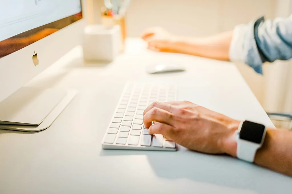 Person typing on Apple iMac keyboard with a smartwatch, in a bright modern workspace.