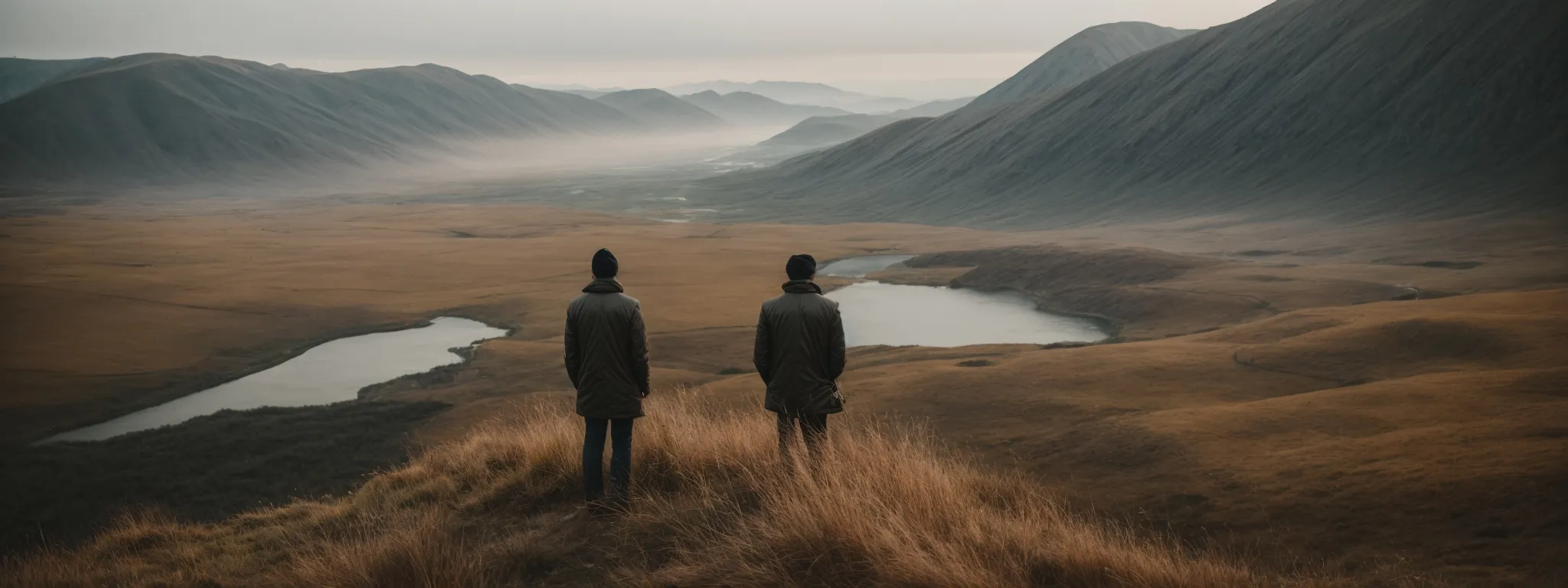 Two people standing on a hill overlooking a valley with lakes and surrounding mountains