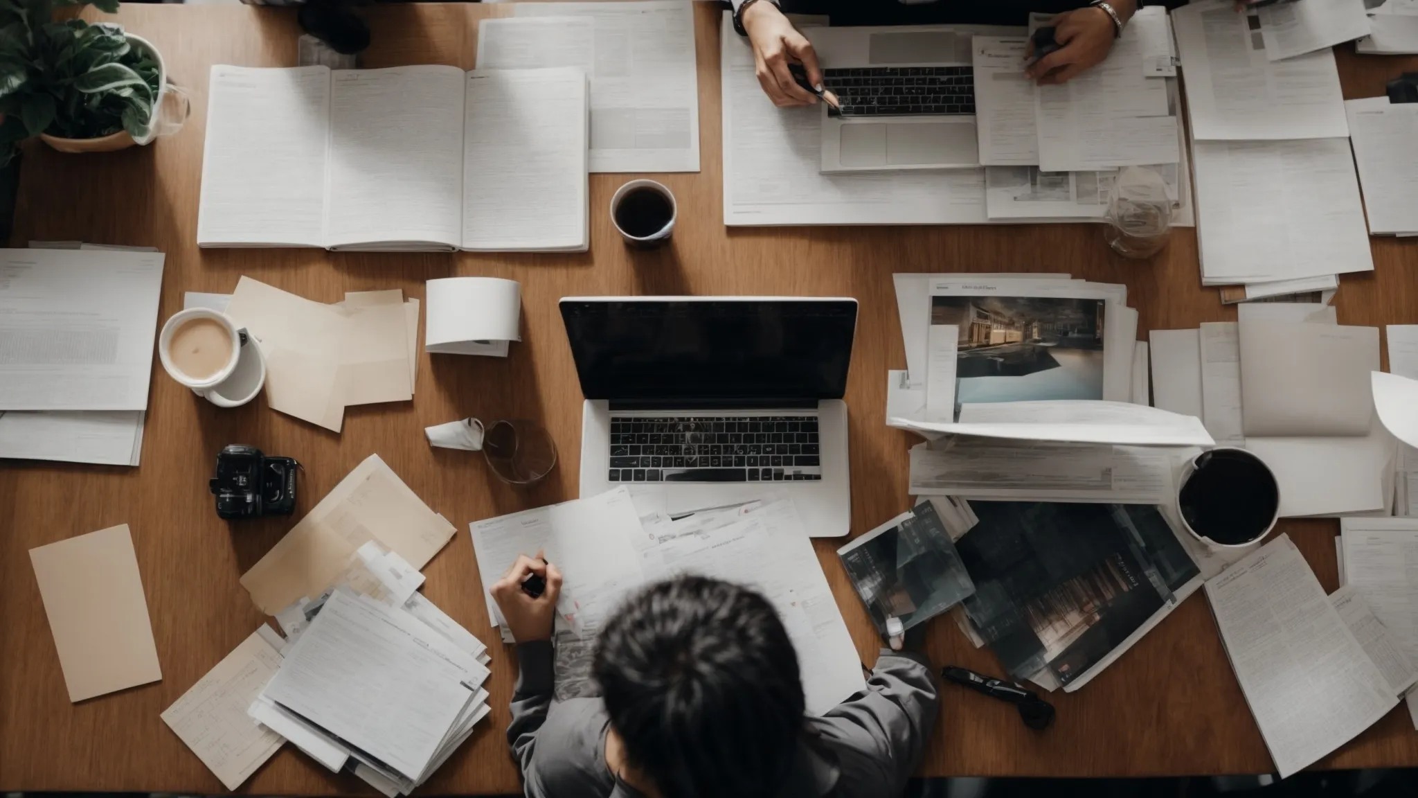 two people sitting at a table with laptops, one surrounded by magazines and the other with analytical charts on the screen.