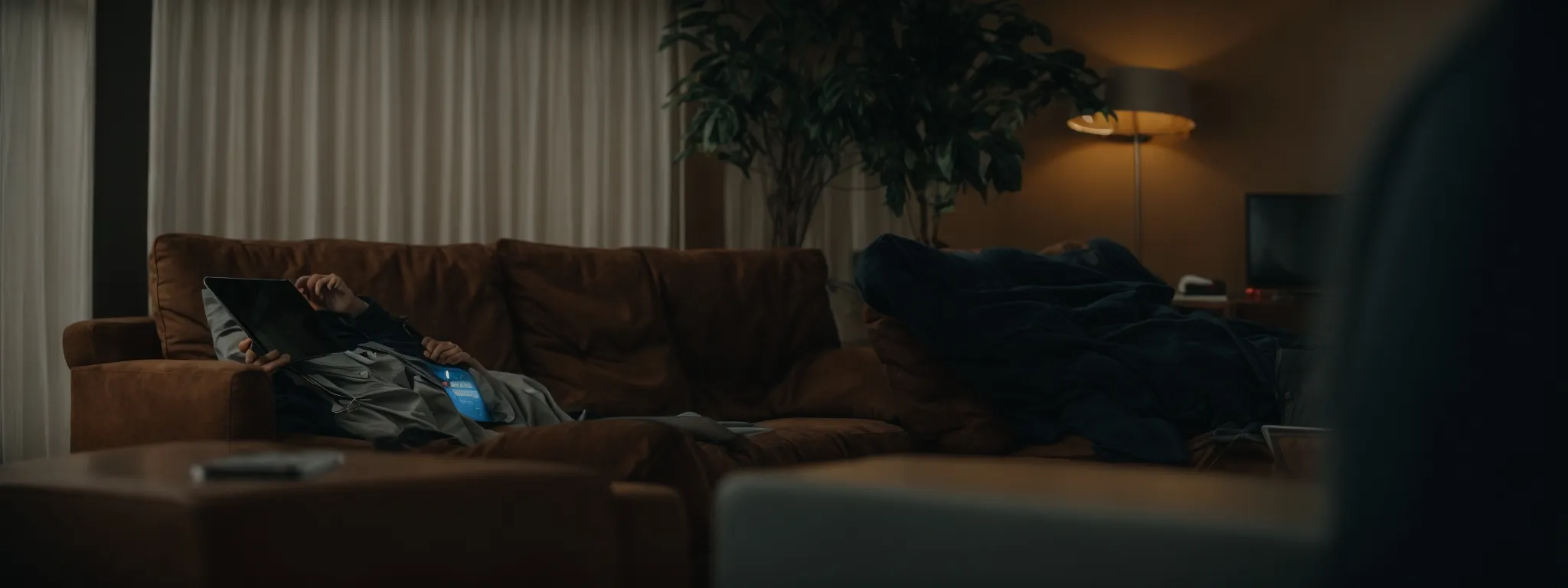 a person lounging on a sofa while using a tablet, with a paused commercial visible on a tv screen in the background.